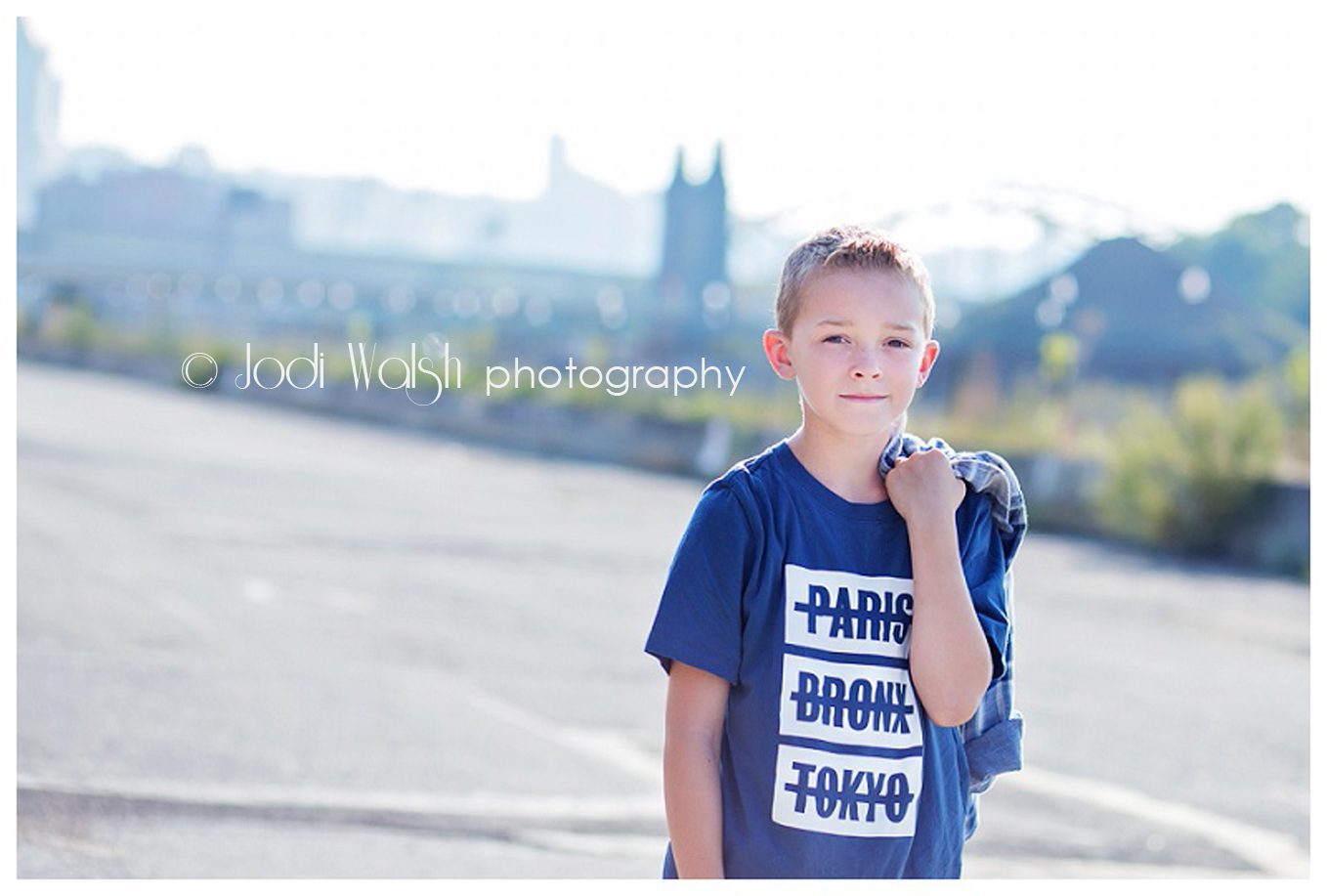 image of a boy wearing a dark blue t-shirt with the words Paris, Bronx, Tokyo in white and holding jacket over his shoulder.  The Pittsburgh city skyline is hazy in the background.