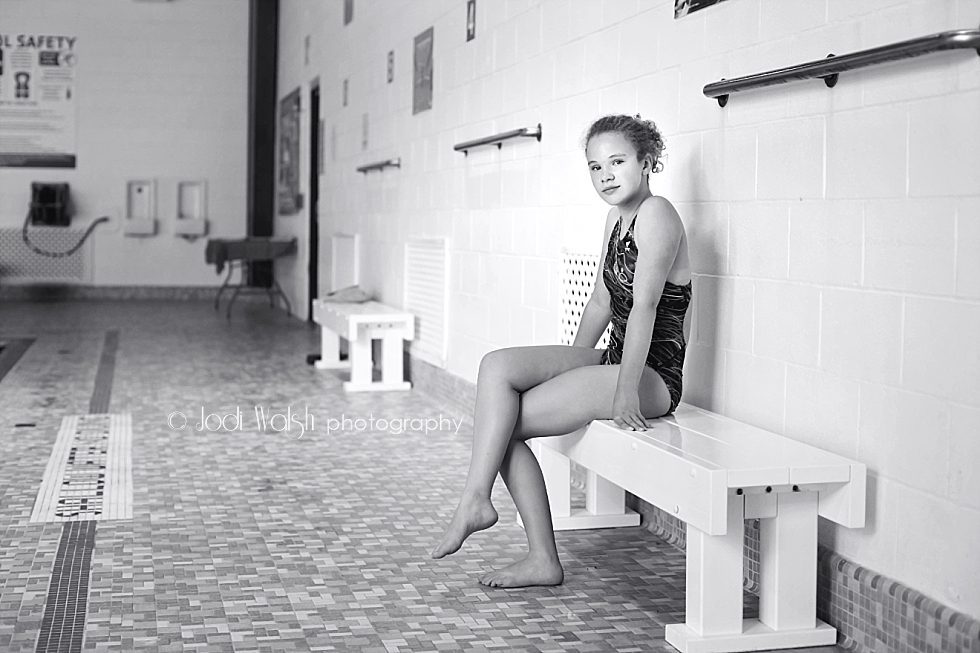 black and white images of a teen swimmer sitting on a bench beside the indoor pool at the Sewickley YMCA. She has curly hair that is pulled back. Her legs are crossed and she's looking thoughtfully at the camera.