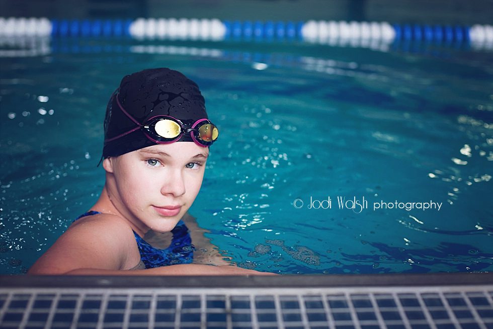 image of a teen girl in the pool. She's wearing a swim cap and goggles as she stands by the edge in the water. She has a thoughtful expression as she looks at the camera.
