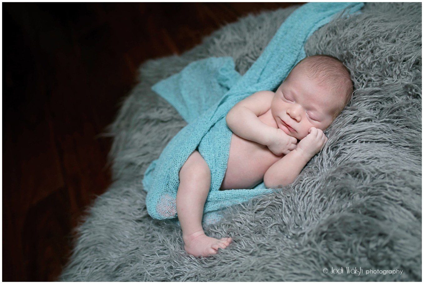 image of a newborn wrapped in a light blue blanket, laying on a gray fuzzy blanket.  He's sleeping with his hands up by his face.