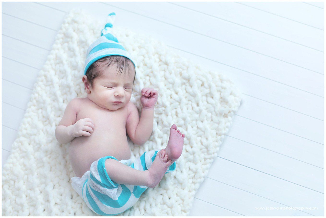 image of a baby boy with blue striped hat and pants. He's sleeping on a cream knitted blanket and white plank floor.