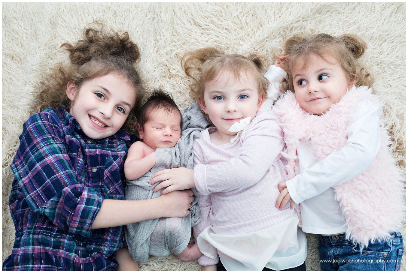 image of three sisters snuggling with their baby brother on a cream fur blanket. The baby is sleeping. The two sisters next to the baby are smiling at the camera. The remaining sister is looking off to the side.