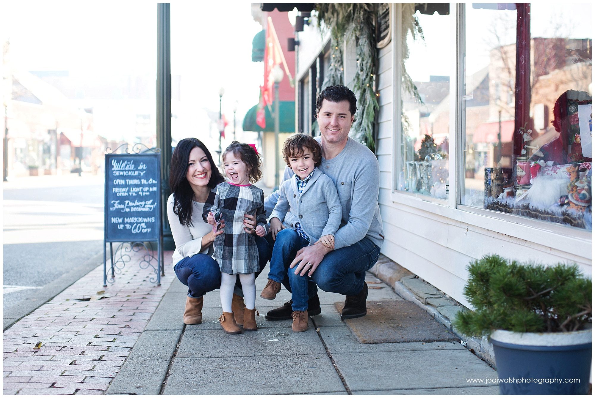 image of a family (mom, dad, little boy, toddler girl). They're kneeling in the middle of a sidewalk in Sewickley. There are Christmas greens in decorating the shops.