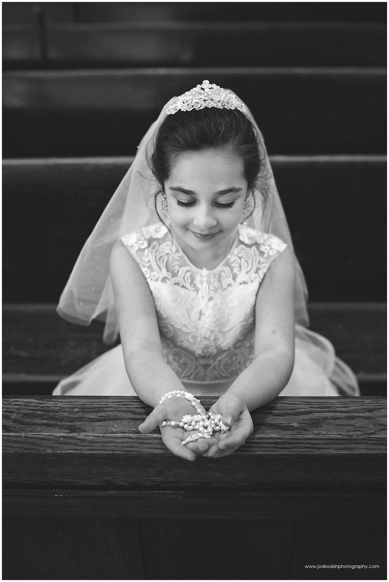 black and white image of a little girl wearing a white first communion dress and veil.  She's sitting in a pew with her hands on the railing, holding a white set of rosary beads. 