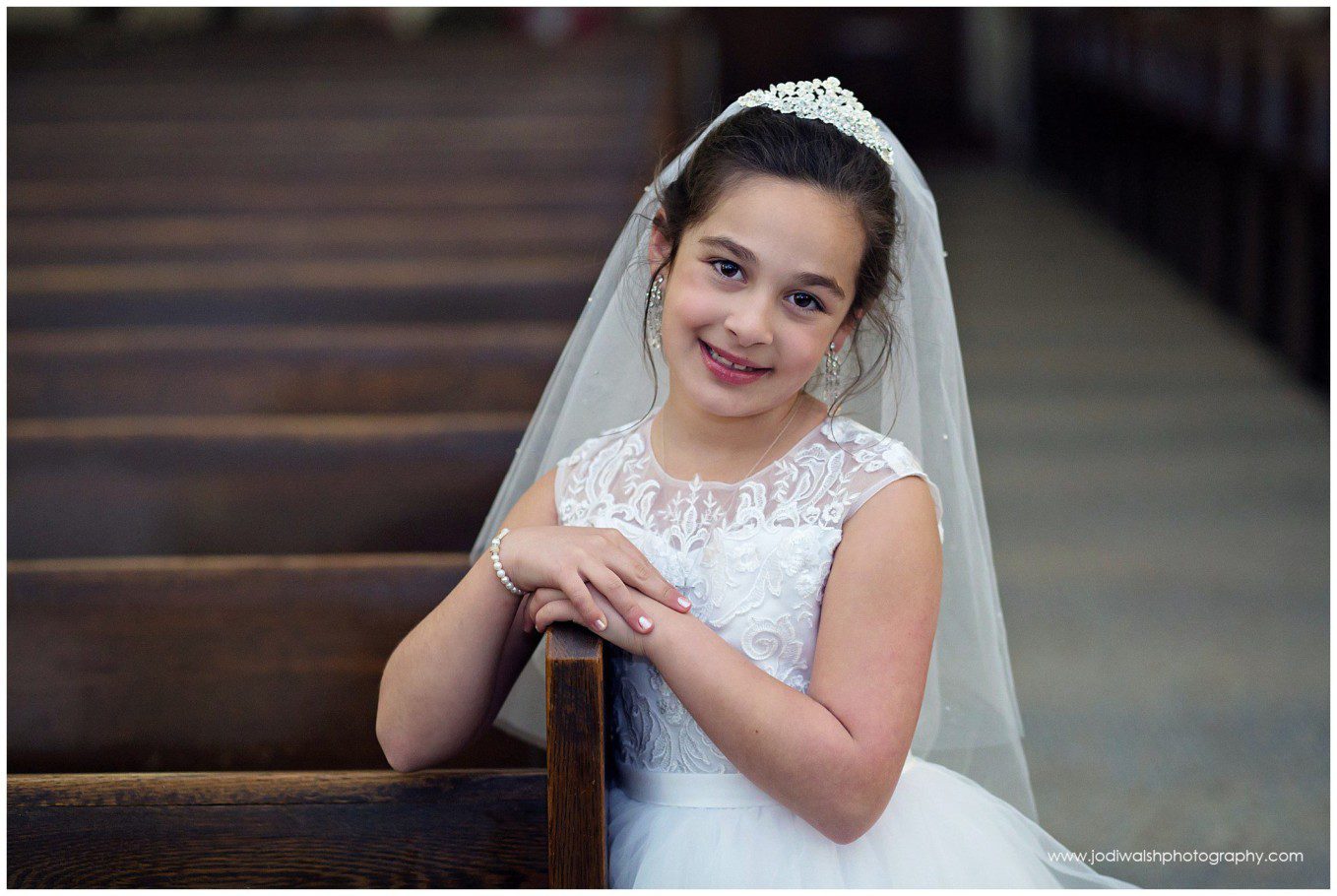 image of a little girl in first holy communion dress, standing next to church pew.  She's looking at the camera and smiling.