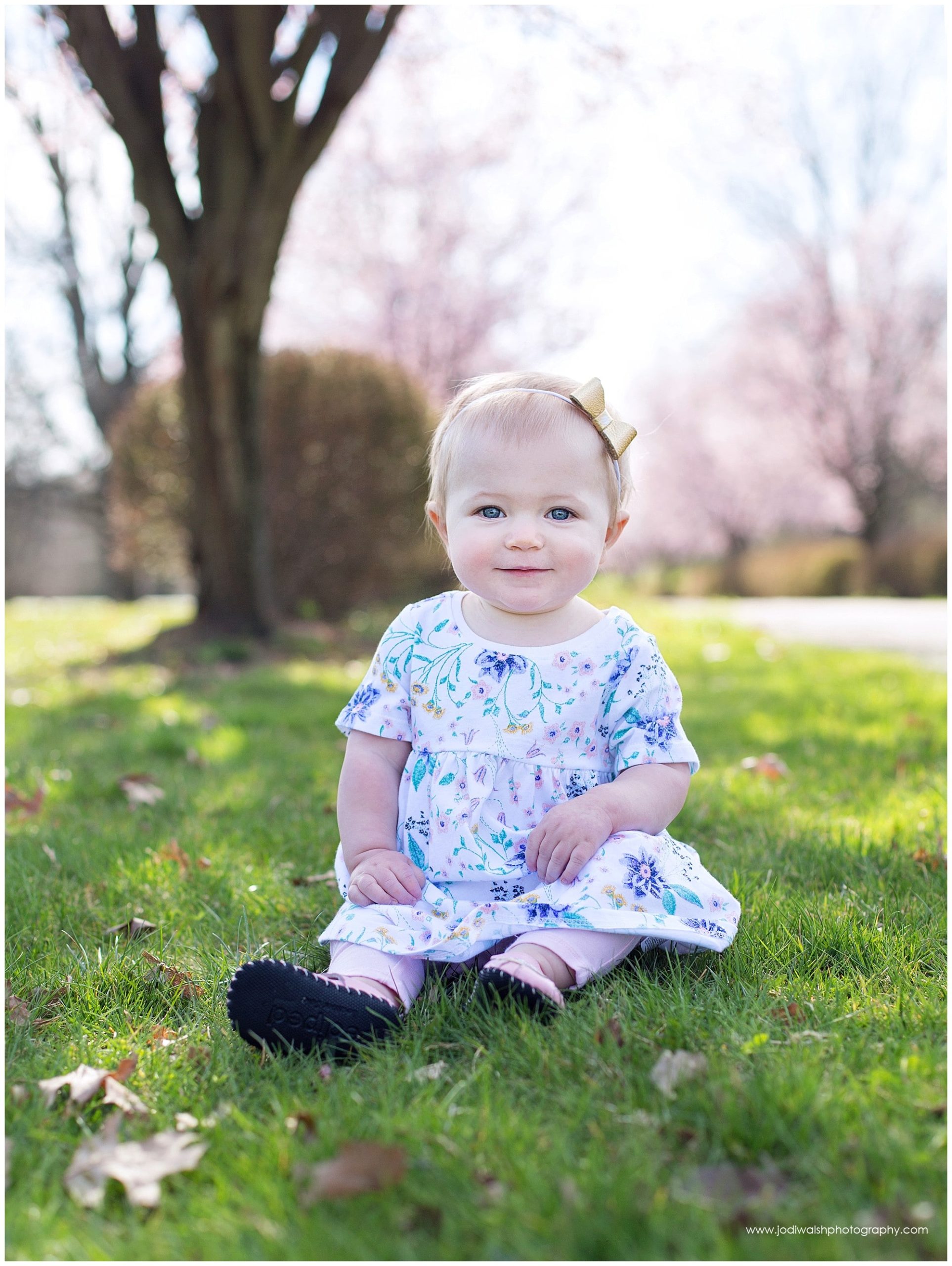 1 year old baby girl sitting in the grass with pink trees behind her