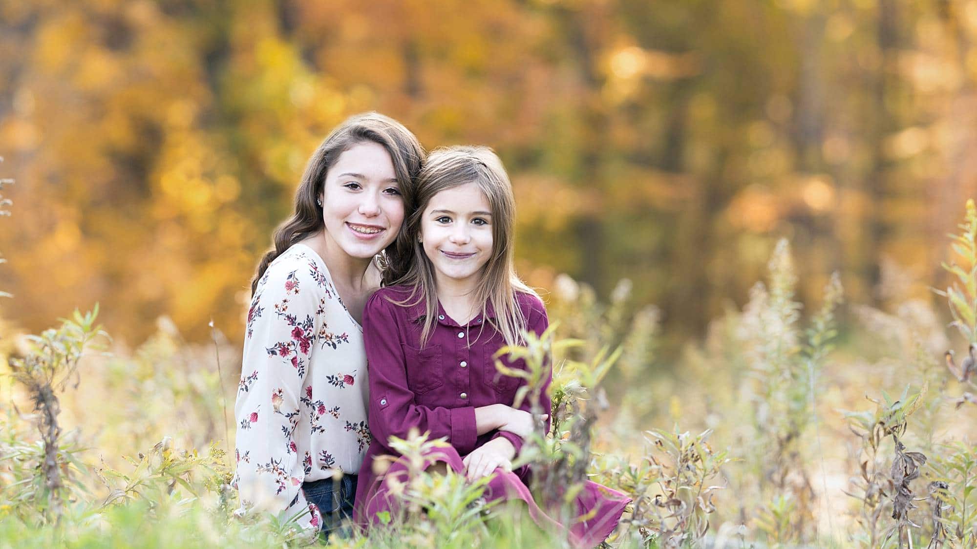 image of sisters sitting in tall grasses. Older teen sister with younger sister in her lap. There are gold trees behind them.