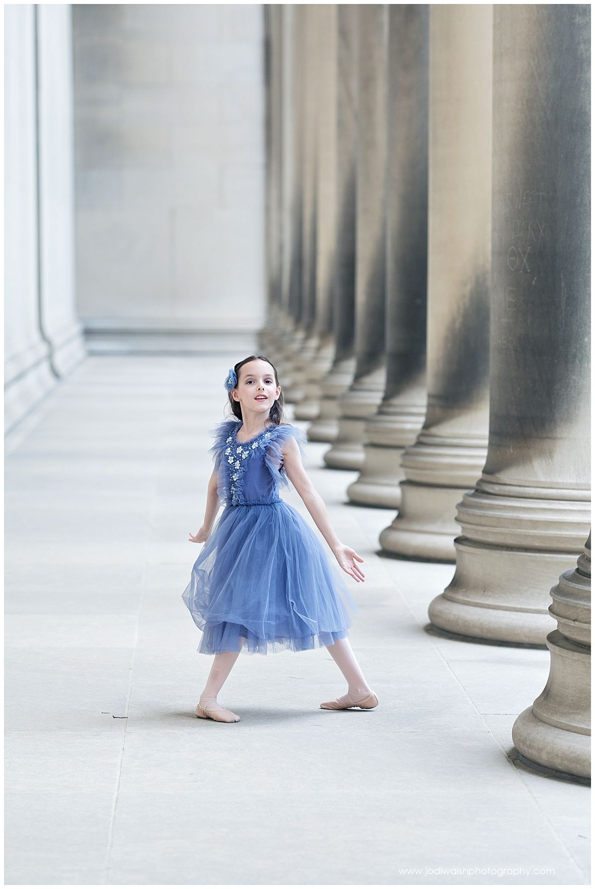 image of a young dancer with long dark hair, wearing a blue tulle dress, posed in a long hallway with stone columns