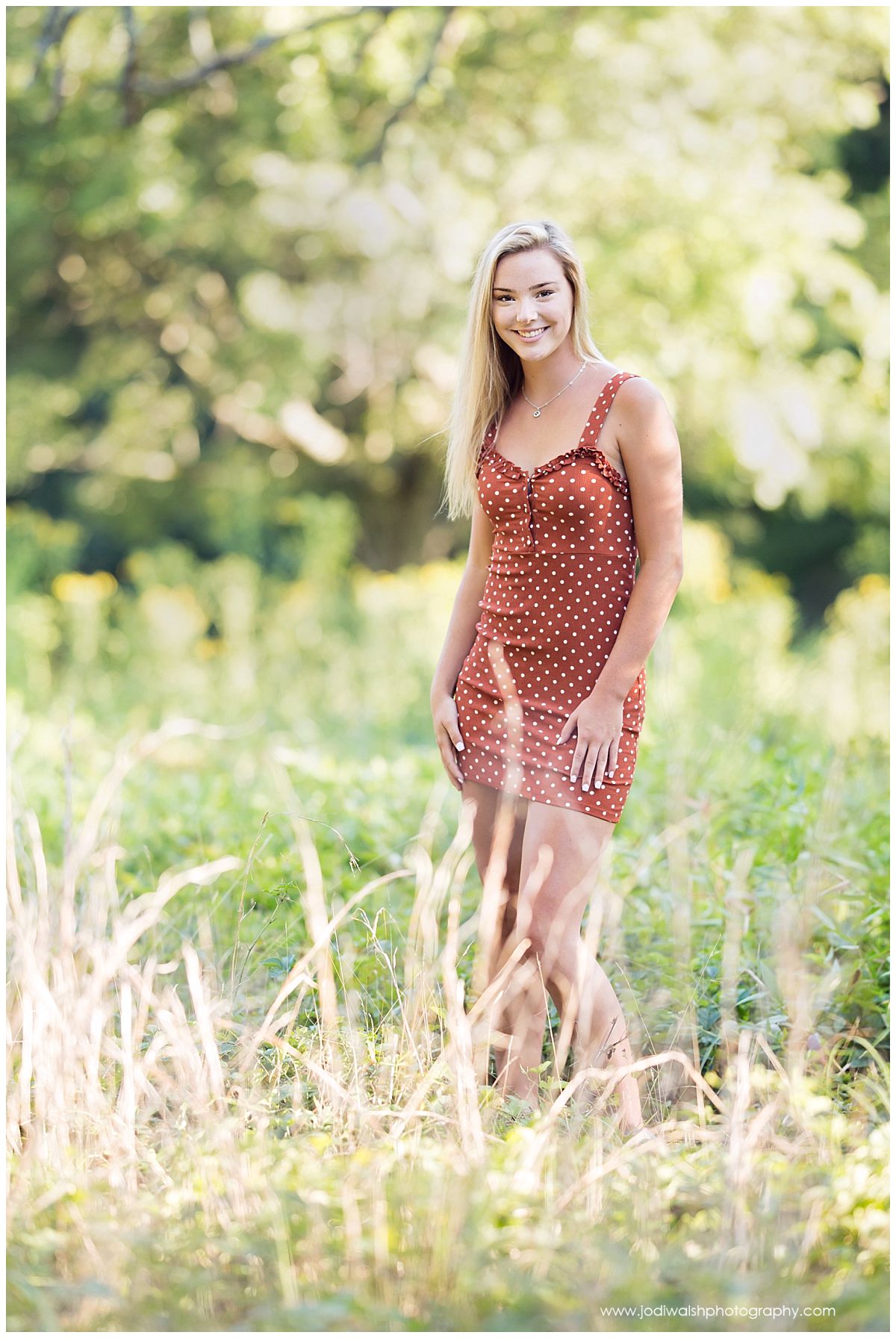 Image of a senior girl in North Park. She's wearing a rust colored polka dot dress and standing in the tall grass, smiling.