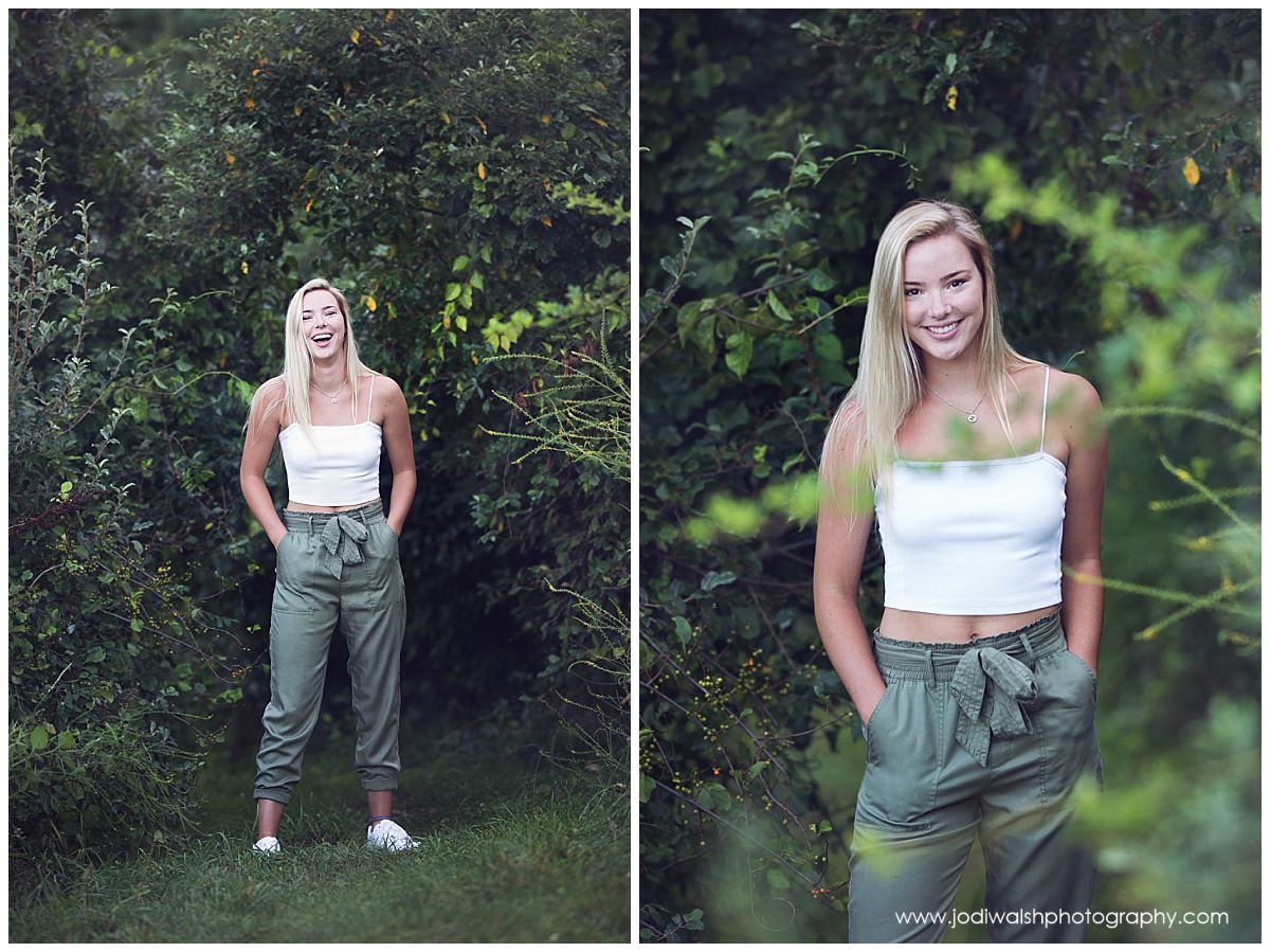 images from a senior portrait session in North Park. Senior girl is standing in a area with tall dark green bushes. She's wearing a white tank top and green cargo pants.