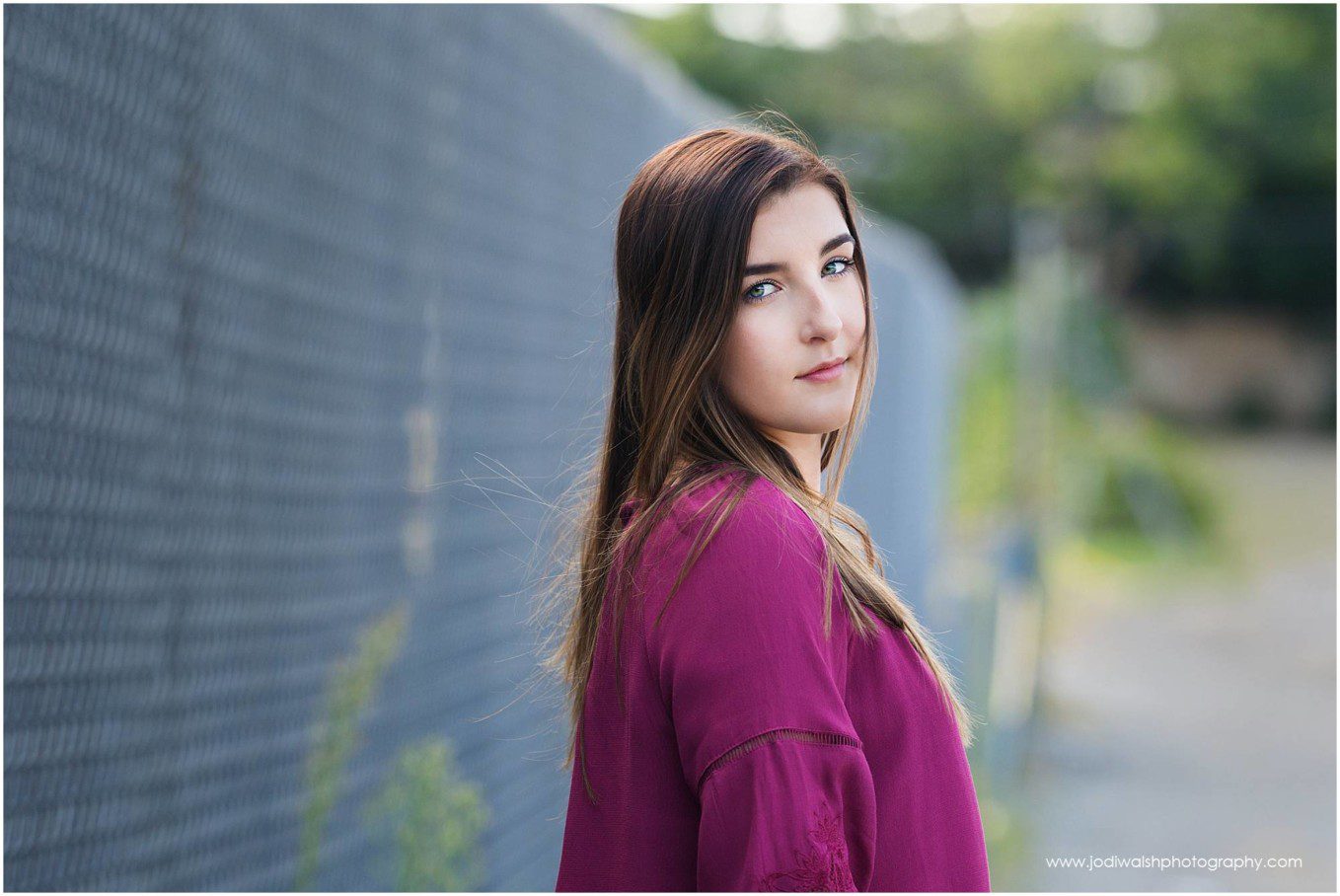 image of a senior girl with dark hair wearing a magenta shirt looking back over her right shoulder. example of Senior portrait image with Jodi Walsh Photography