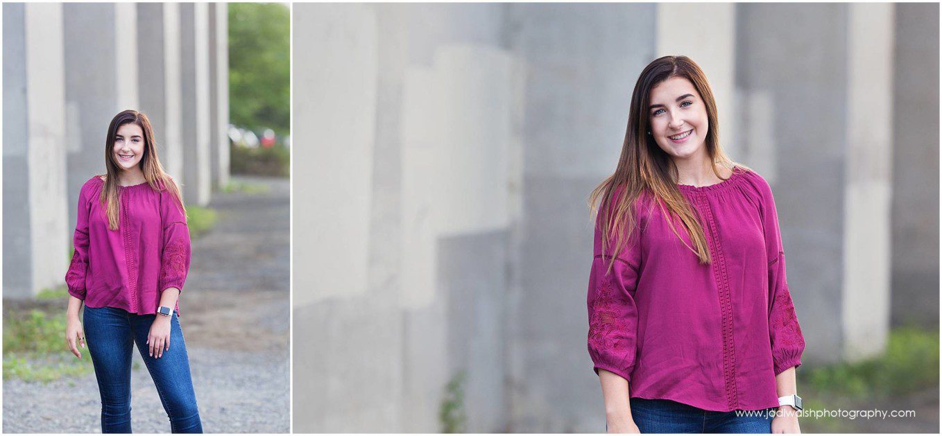 image of a senior portrait session along the North Shore Trail in Pittsburgh. The senior girl is wearing magenta boho top and jeans while standing next to a gray cement wall.