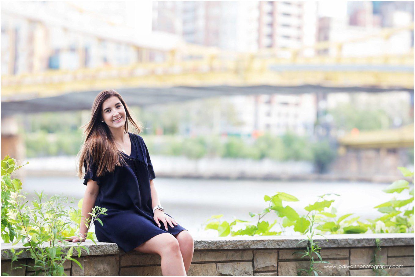 Image of a senior girl wearing a black dress sitting on a stone wall along the North Shore Trail, Pittsburgh. 