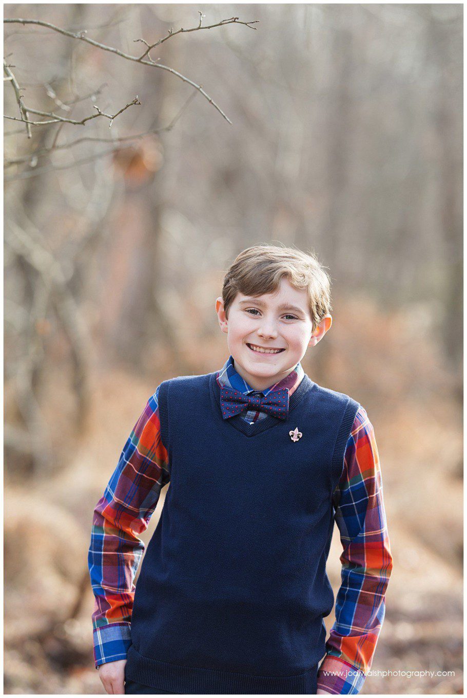 Image of a teen boy wearing a navy sweater vest and bow tie. He's standing in a winter woods with bare trees and tall grasses.