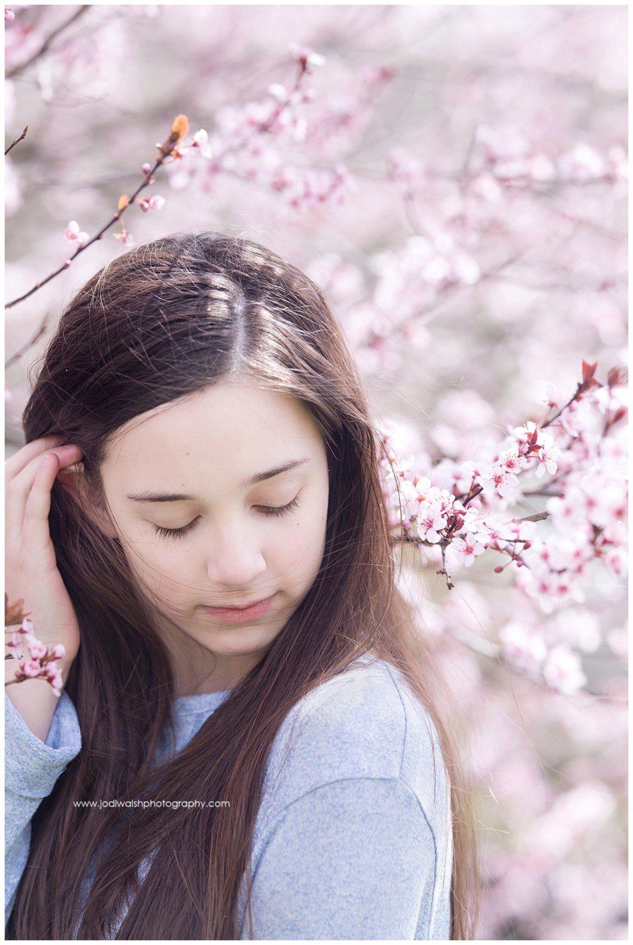 image backyard spring portrait with Jodi Walsh Photography. the photo is of a teen girl with dark hair with pink blossoms all around her. She's looking down at her shoulder and gently pushing her hair behind her ear.