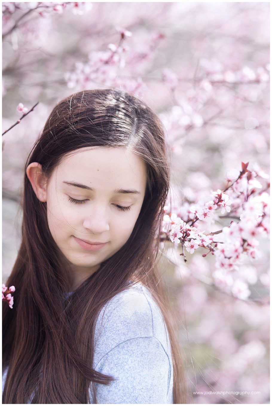 image backyard spring portrait with Jodi Walsh Photography. the photo is of a teen girl with dark hair with pink blossoms all around her. She's looking down at her shoulder.