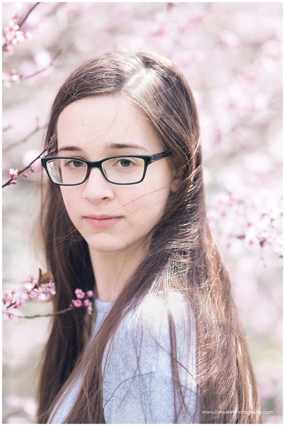 images of a teen girl standing near a tree with light pink blossoms. She's looking over her shoulder, wearing glasses and has a thoughtful expression