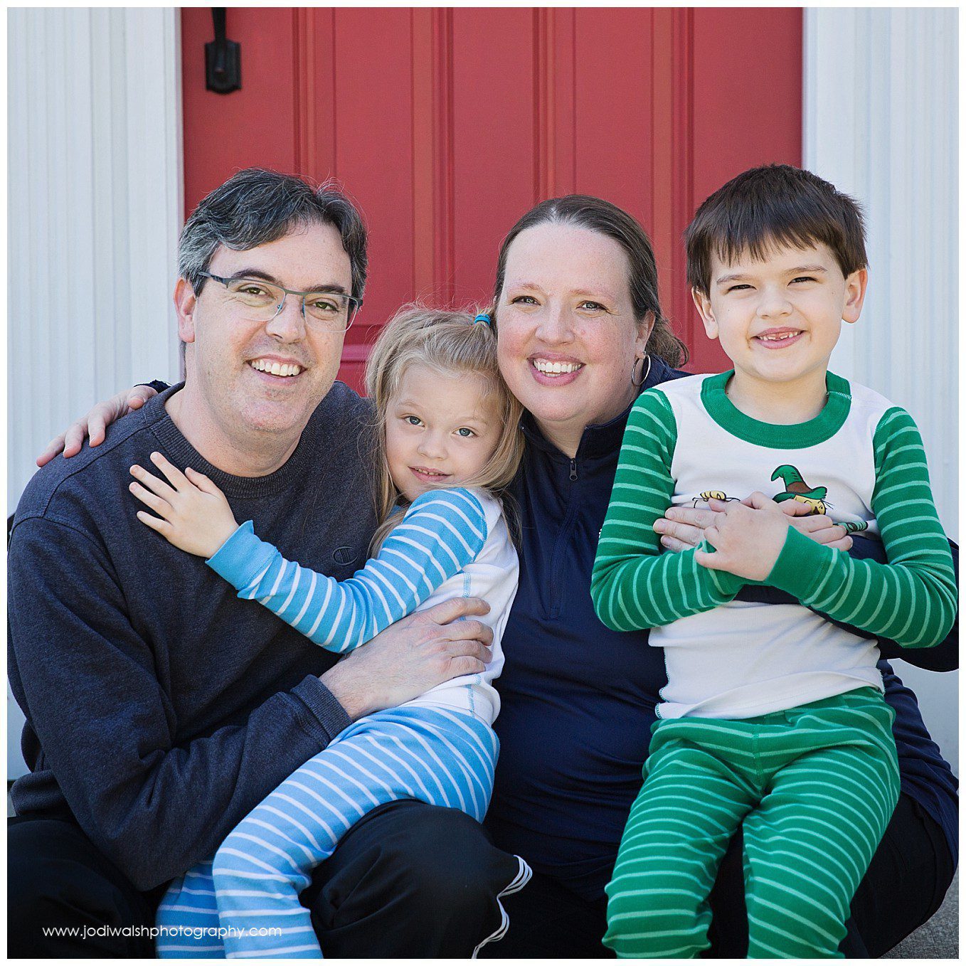 image of a family sitting together on their front porch. Dad has a little girl sitting in his lap and she's wearing blue striped pajamas. Mom has a little boy sitting in her lap and he's wearing green striped pajamas. There's a red front door behind them. This is a sample of a porch portrait taken by Jodi Walsh Photography during the 2020 pandemic.