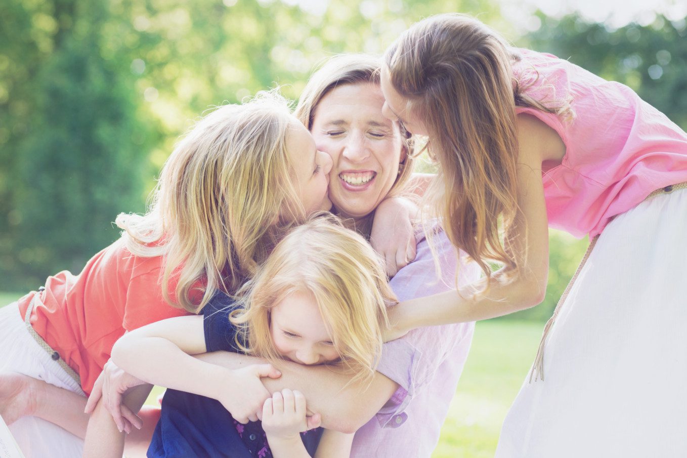 Image of a mother with three young daughters.  Mom is in the center with her girls all covering her with kisses. They're all grinning.  We celebrate moms with kisses.