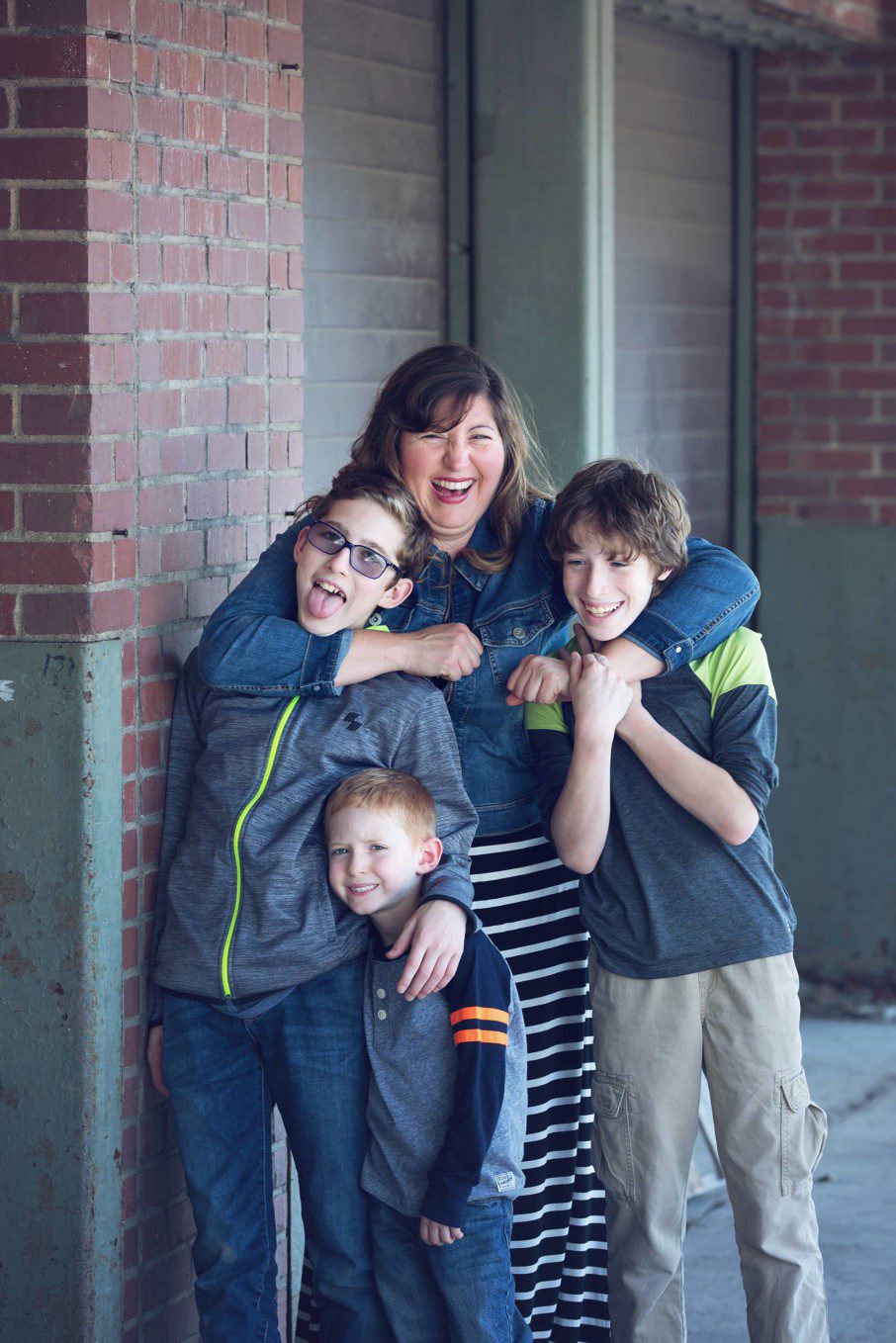 celebrate moms of boys.  Image of mother with three sons standing next to a brick wall in the Strip District..  Two of the boys are teens and mom has her arms around their necks.  The littlest boy is hugging his older brother.  They're are all grinning.  