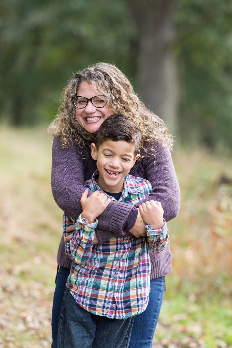 image of a mother hugging her young son.  The boy is in front and grinning with his eyes closed.  Mom is standing behind with her arms wrapped around his shoulders.  She's looking at the camera, smiling.  They're in a park in the fall.