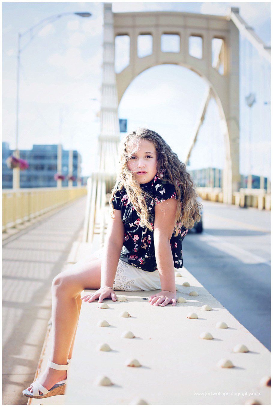 Image of a teen portrait by Jodi Walsh Photography. the image shows a teen girl with curly hair, sitting on the yellow metal beam of the Rachel Carson Bridge in Pittsburgh. The wind is blowing her hair slightly and she has a serious look.