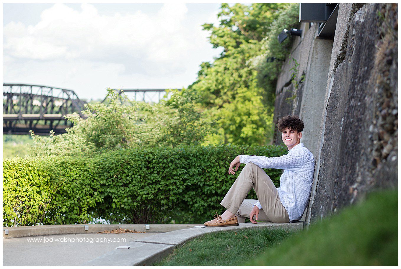 image of a senior portrait by Jodi Walsh Photography. image shows a senior young man in a white dress shirt and khaki pants, sitting on a stone wall, smiling. You can see an outline of the Hot Metal Bridge in the distance.