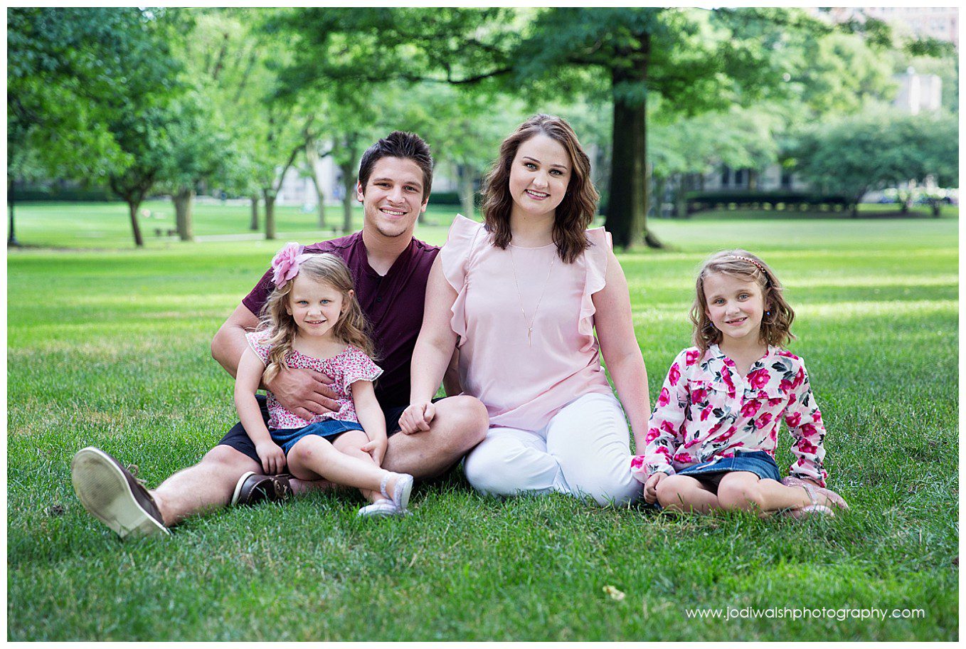 image of a family portrait by Jodi Walsh Photography. the image shows a family with mom, dad, and two young girls sitting on the lawn at the Cathedral of Learning, Pitt campus, Pittsburgh