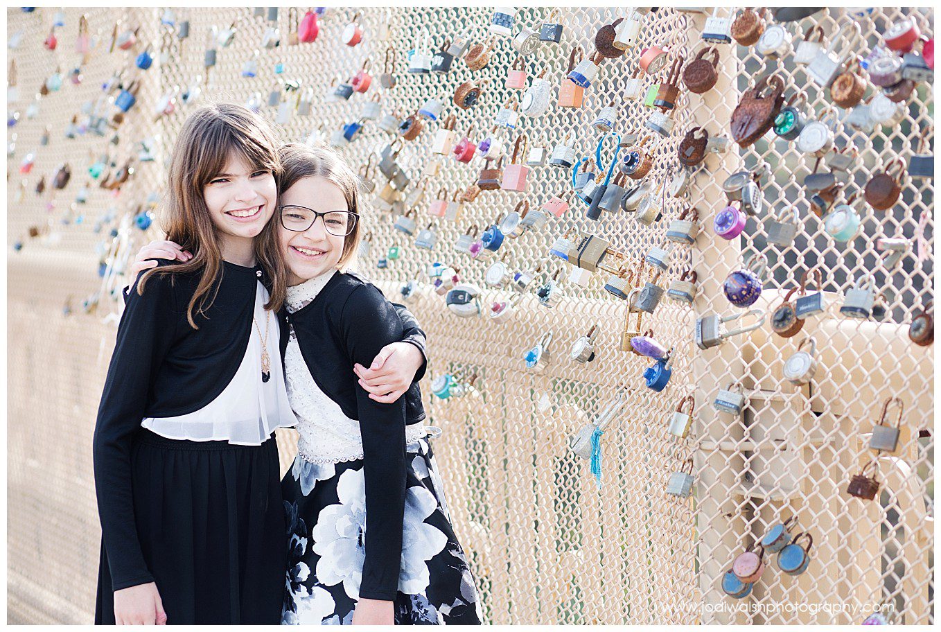 image of pre-teen sisters hugging on a bridge in Oakland. The younger sister wears glasses. They're both smiling. The bridge fencing is covered with a variety of combination locks and padlocks.