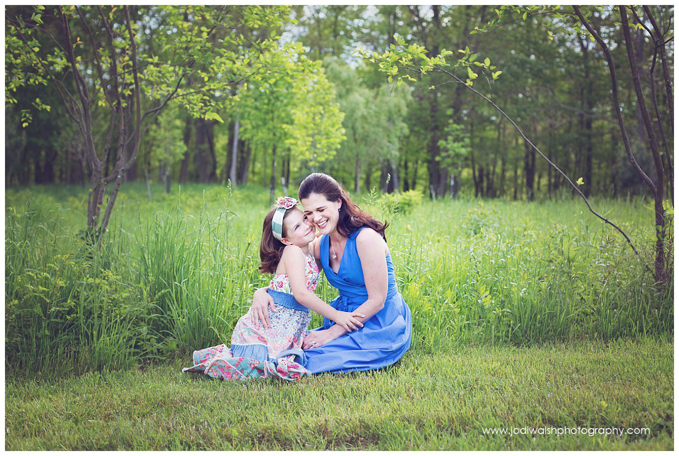 image of a mother and daughter sitting in the grass in a beautiful green park. The daughter is looking up at her mother and grinning. The mother is looking down and laughing.