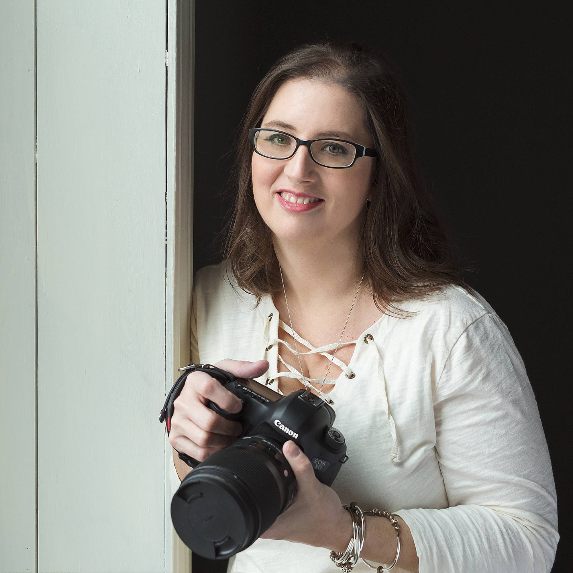 image of photographer, Jodi Walsh. She's leaning next to a doorway, wearing a white top and holding her camera. She's wearing dark frame glasses and is smiling.