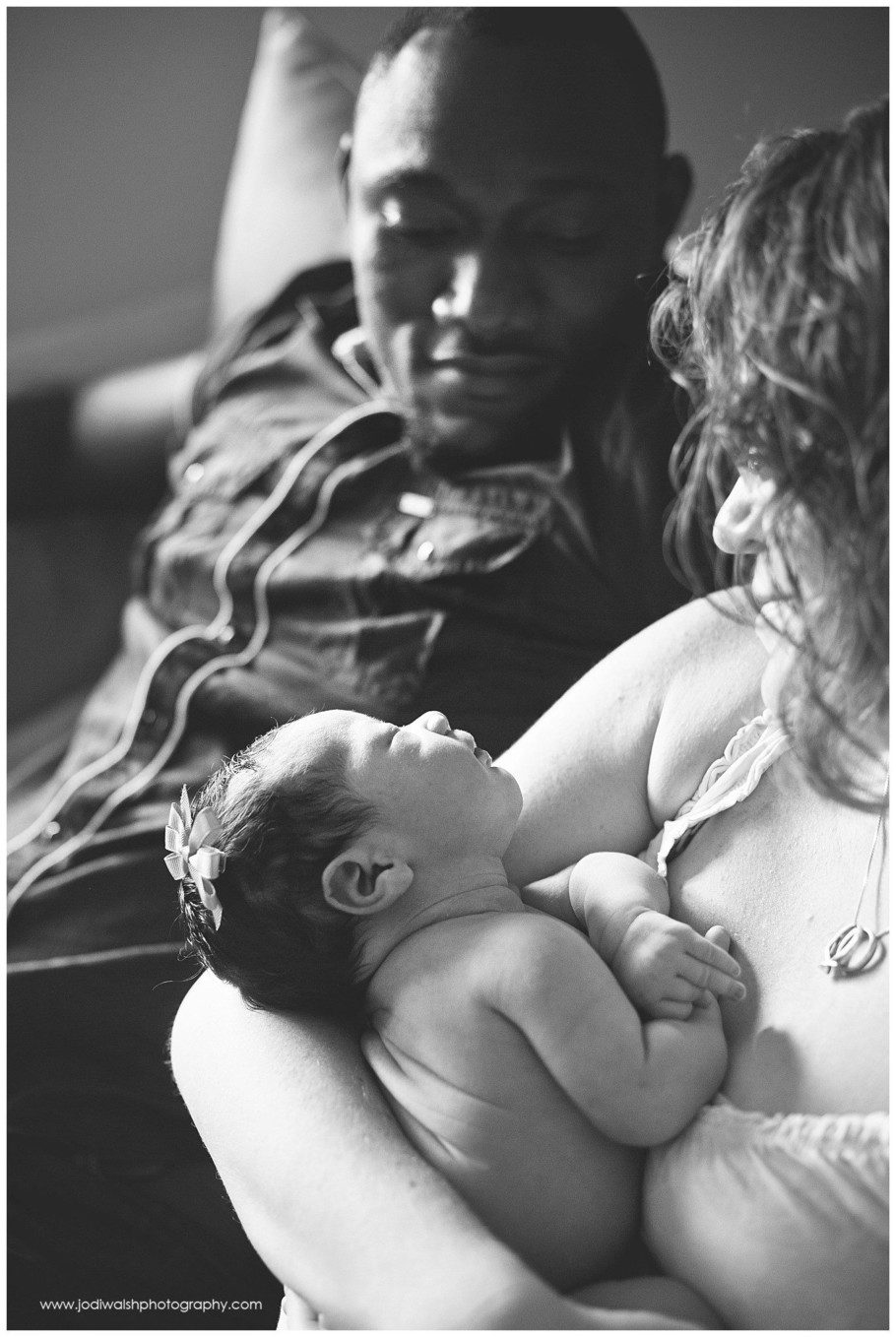 black and white image of a father and mother and newborn baby girl in their home. The dad is in the background looking intently down into his daughter's face as mom cradles her looking down too.