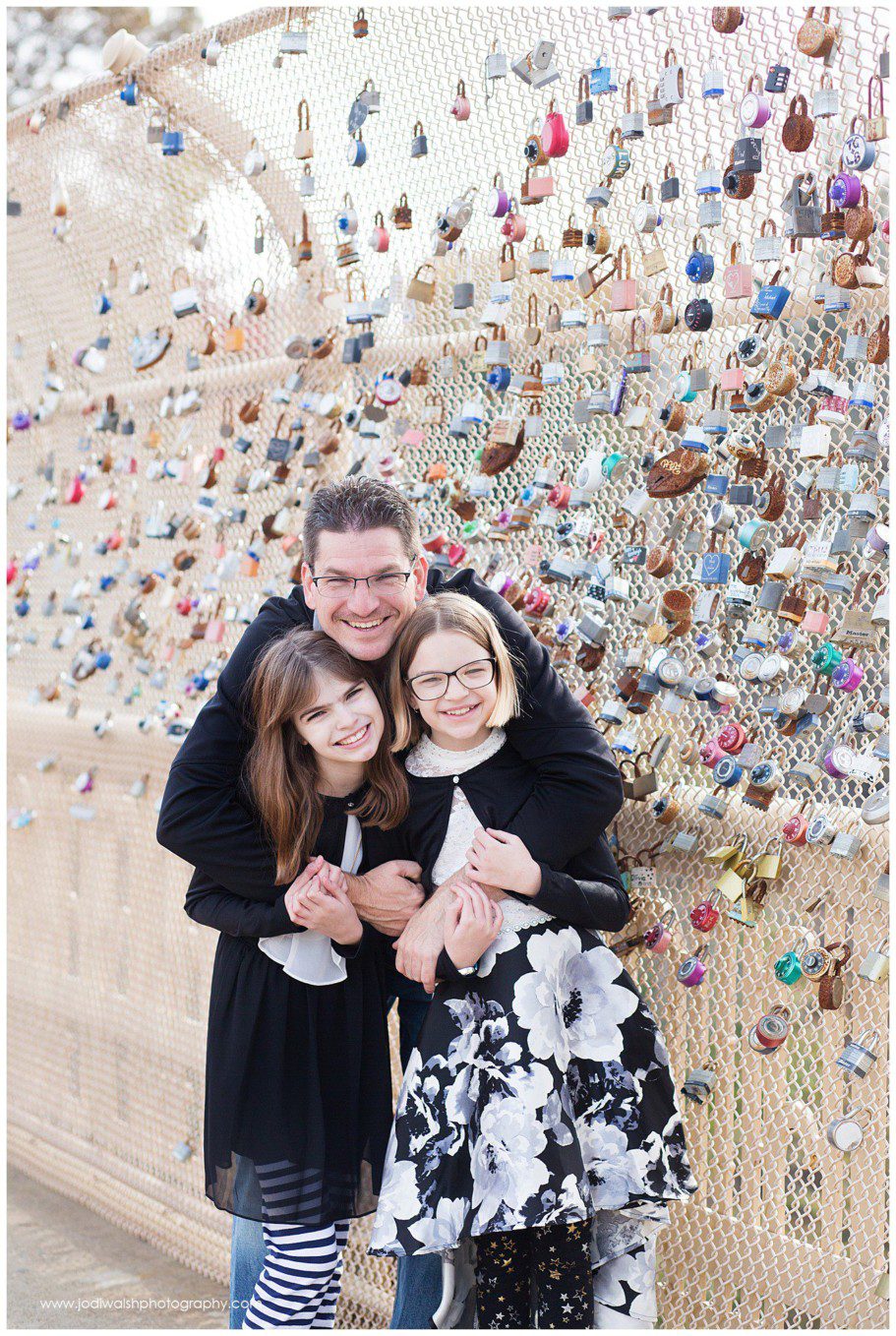 image of a dad hugging his pre-teen daughters while standing on a bridge in Oakland. The fencing on the bridge is covered with different sized and colored pad locks.