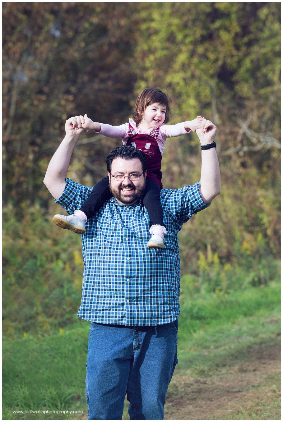 image of a dad with his toddler daughter riding on his shoulders. He's wearing glasses and has a dark beard. They're both grinning and holding hands.