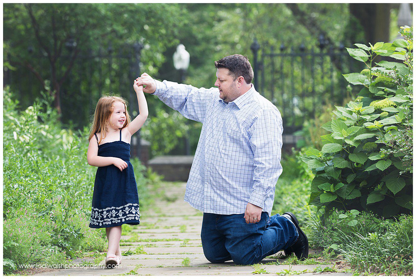 Image of a father and young daughter. Daddy is kneeling and looking at his daughter while holding her hand as she smiles at him and goes to twirl. They're on a pathway in Mellon Park.