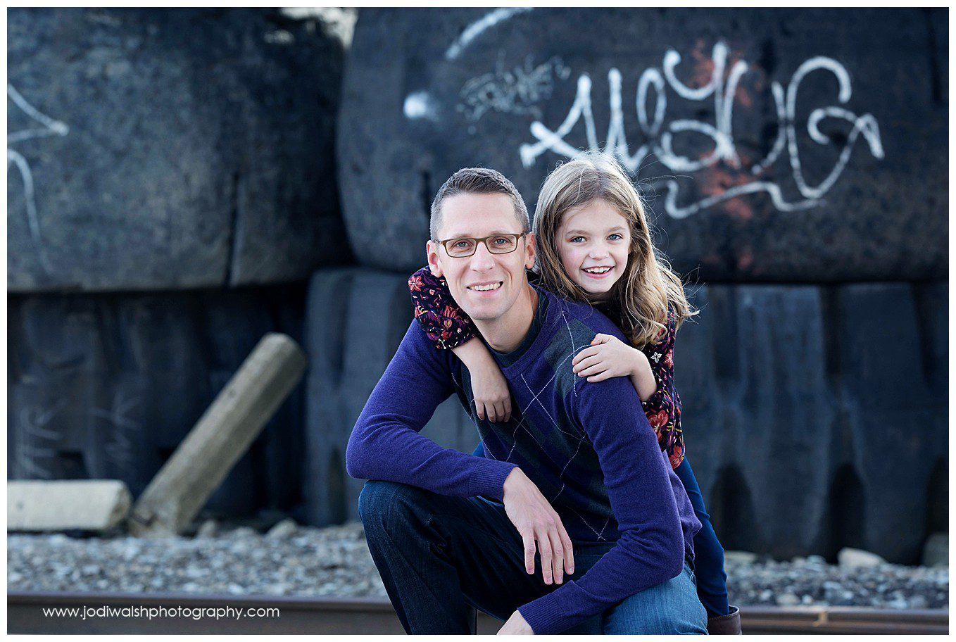 image of a dad kneeling down with his elbows on his knees and his daughter standing behind him, her arms around his shoulders. They're both smiling. They're posed in front of large industrial tires with graffiti on them. 