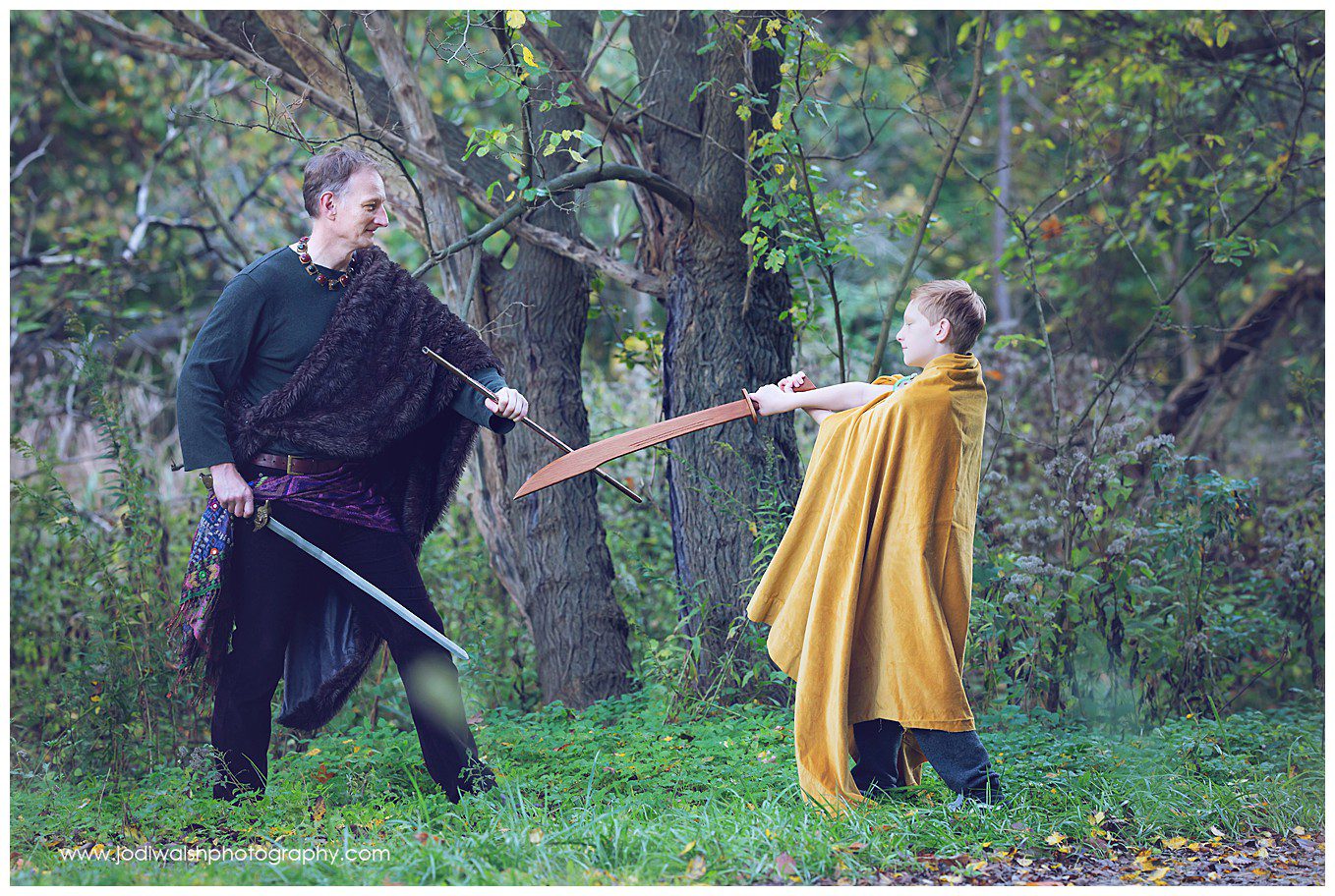 image of a father and son dressed in costumes with capes having a pretend sword fight in the woods of Schenley park. The boy is early teens and is wearing a long gold cape and holding a wooden sword as his dad blocks the strike.