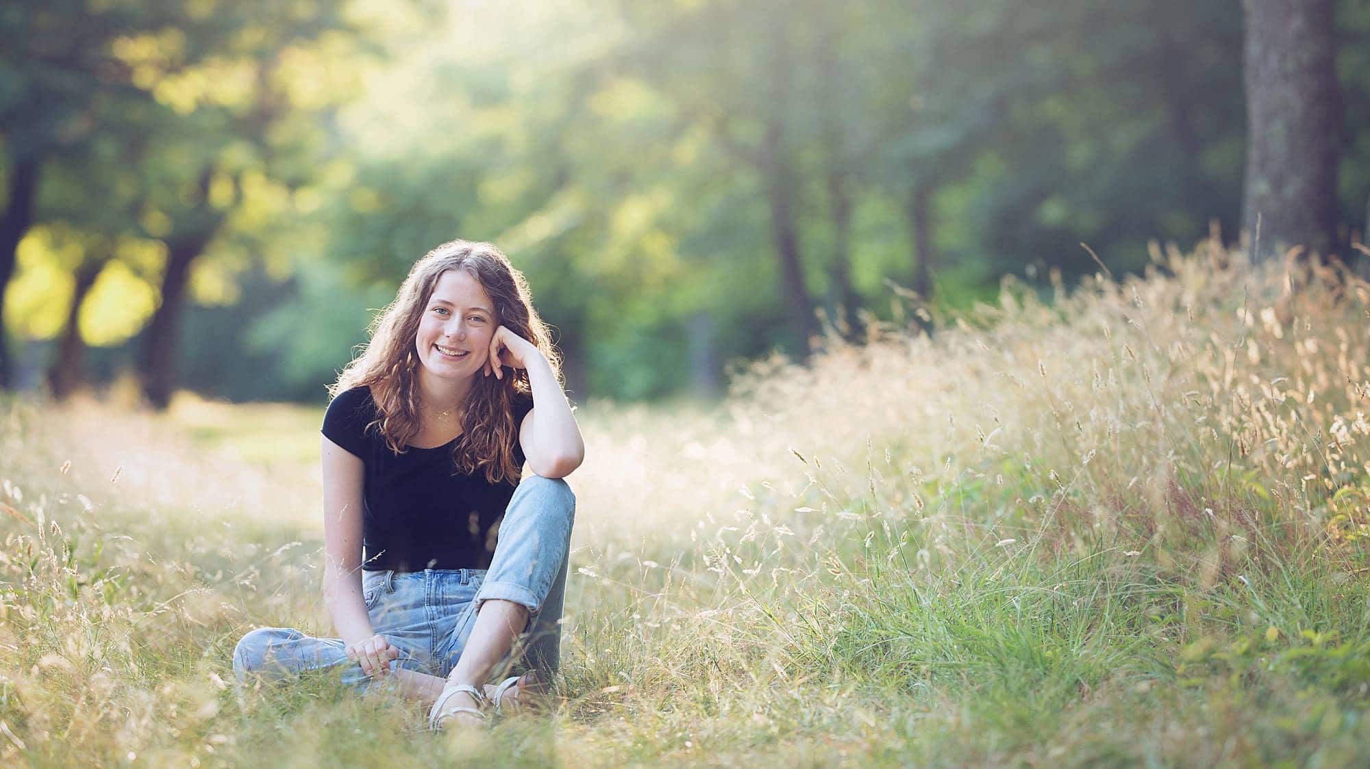 image of a senior girl with dark curly hair, wearing jeans and a black t-shirt, sitting in a grassy field with her elbow on her knee and the sun streaming behind her.