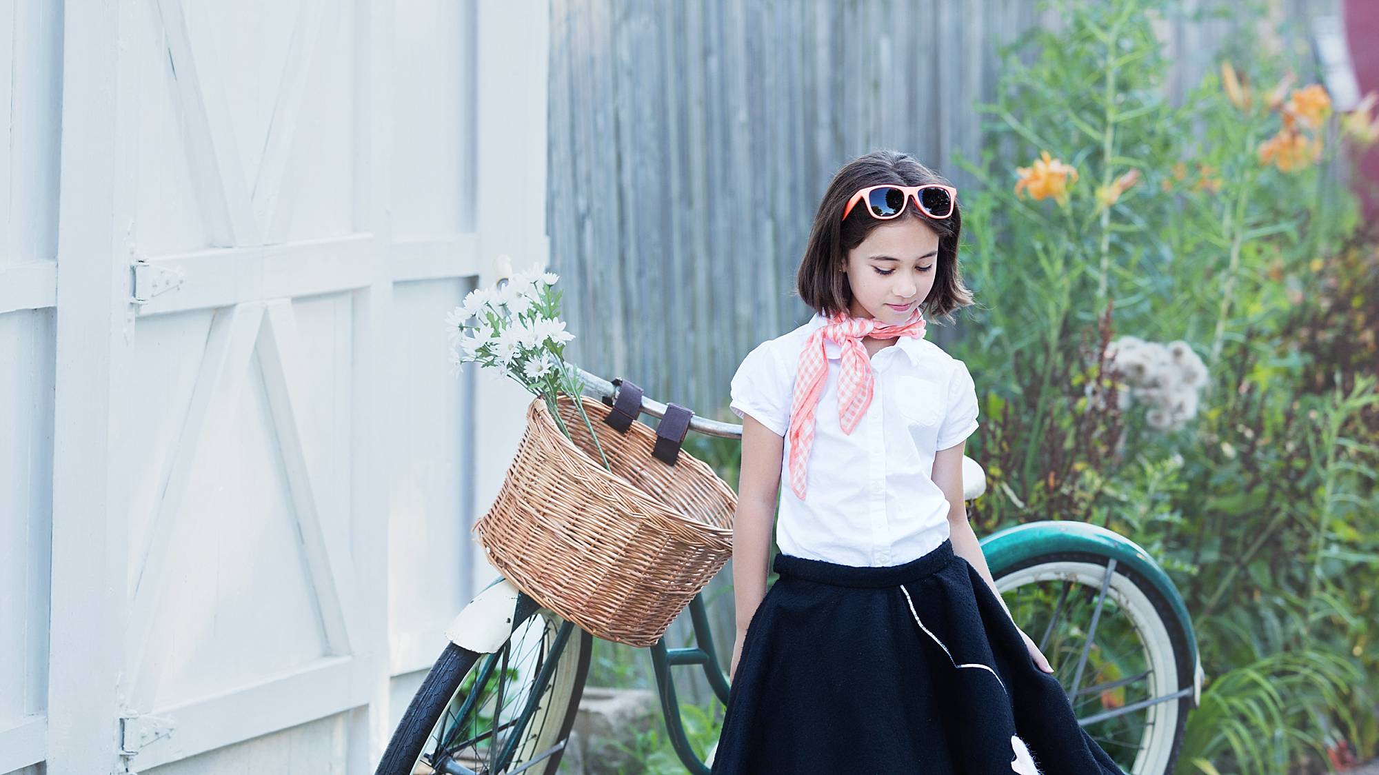 image of a little girl with short dark hair, looking down and standing next to an old fashioned bicycle with a wicker basket and daisies