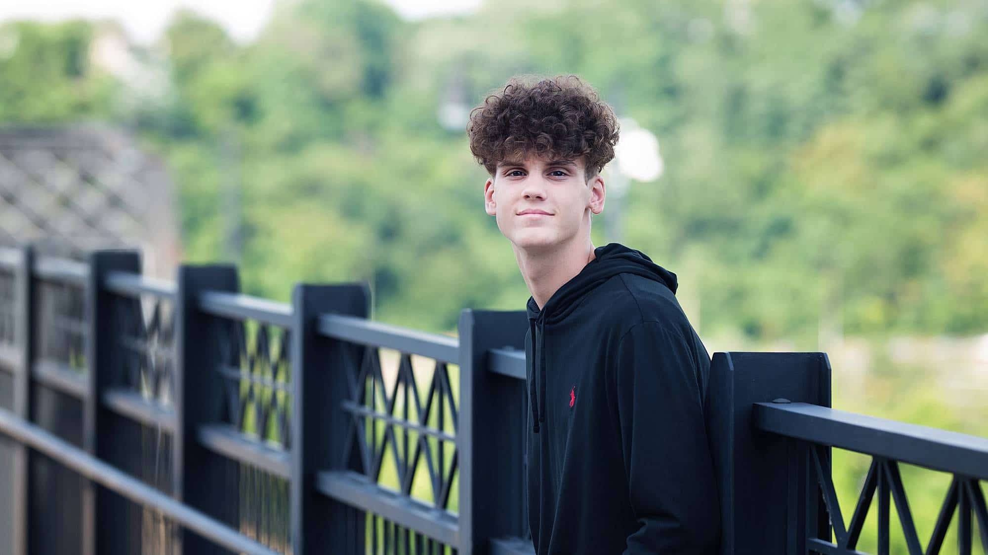 image of a senior guy with dark curly hair, wearing a black hooded sweatshirt, leaning against an black railing