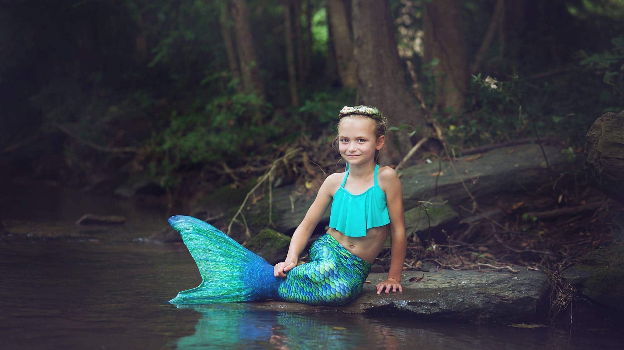image of a little girl, sitting by a creek, dressed in a teal blue mermaid tail