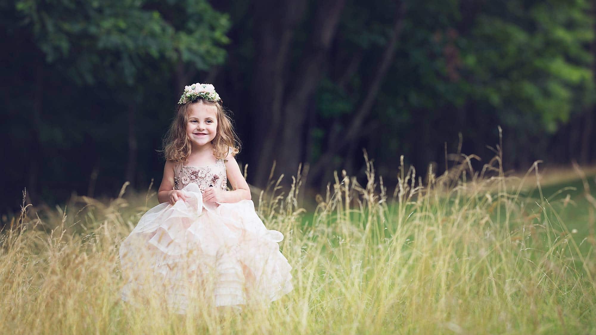 image of a little girl with curls wearing a pink princess dress and flower crown, standing in tall golden grasses