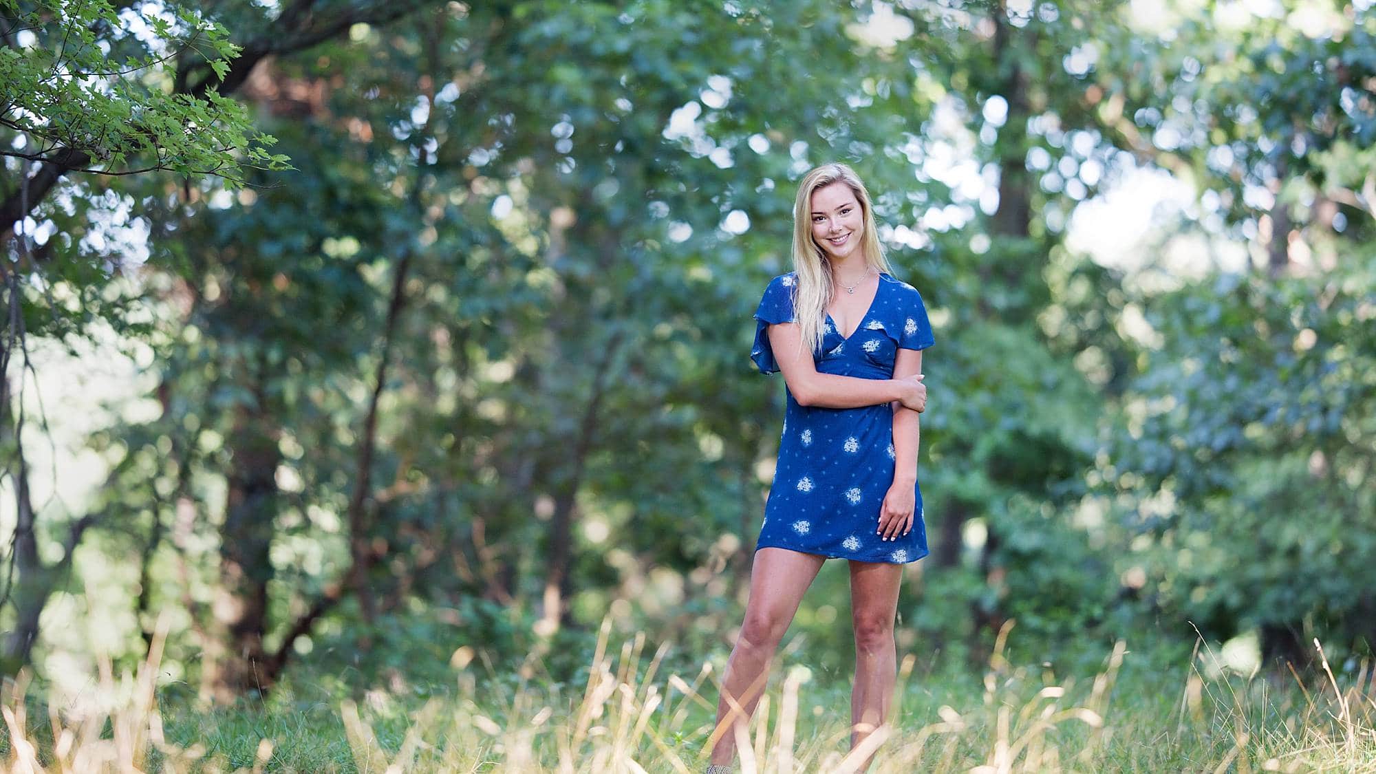 image of senior girl with blonde hair wearing a short blue dress with white flowers and standing in a wooded and grassy field