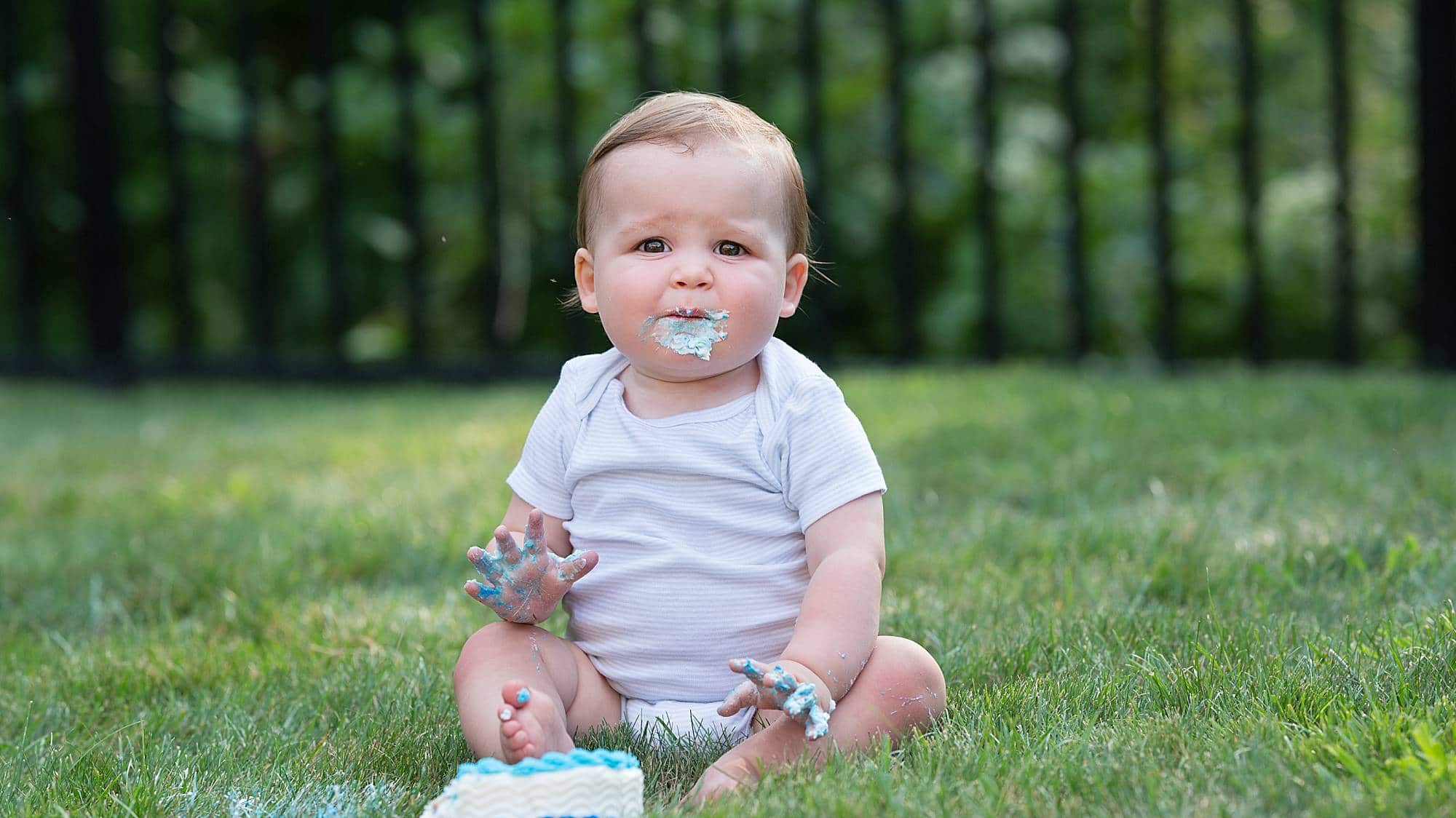 image of a baby boy with frosting on his chin sitting in a grassy yard with a birthday cake
