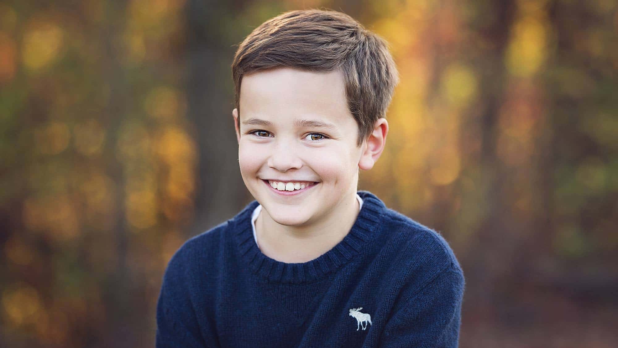 image of a young boy grinning, wearing a navy blue sweater with fall trees in the background