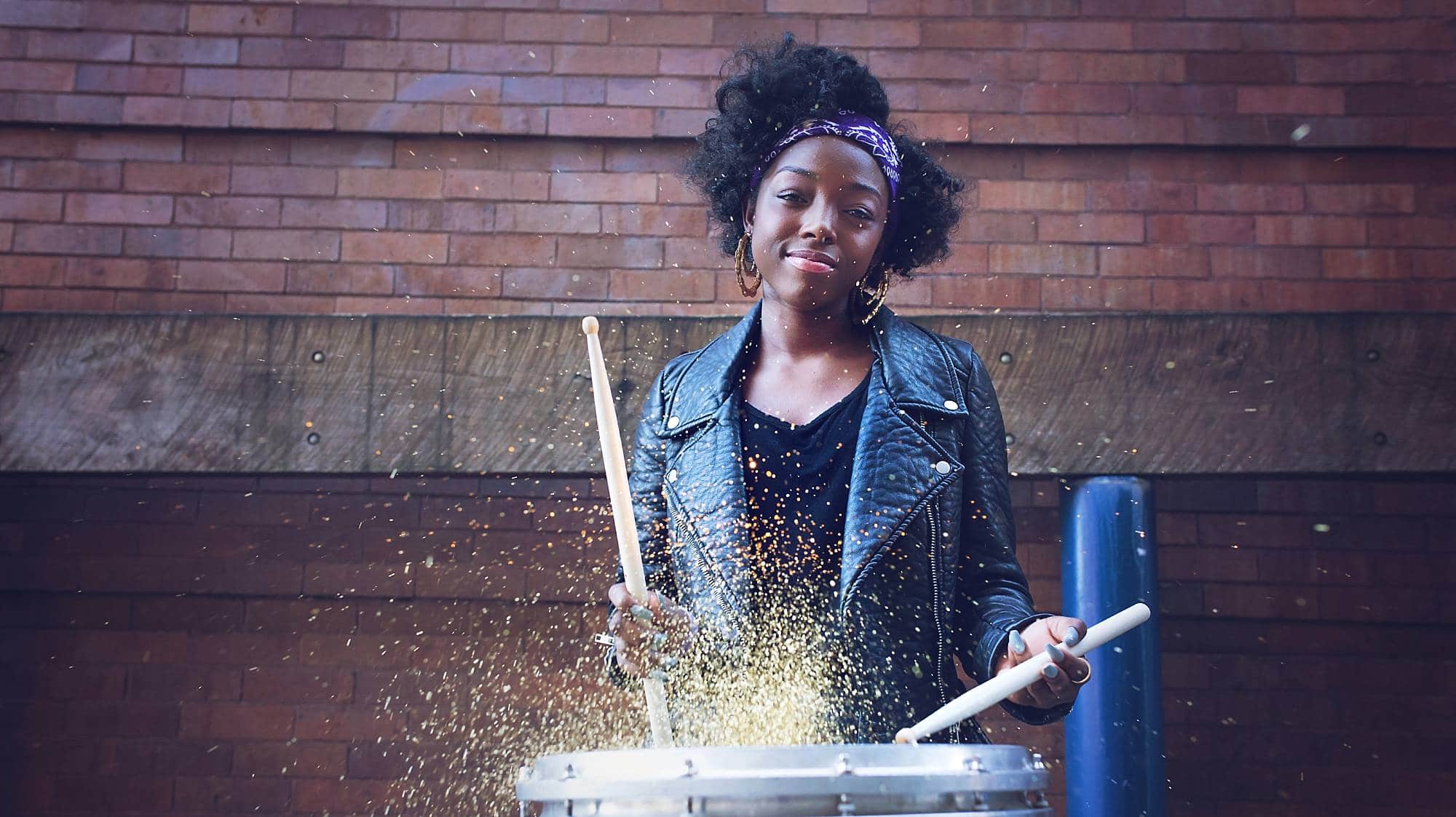 image of a black girl in a leather jacket and black top, hitting a snare drum with gold glitter puffing up.