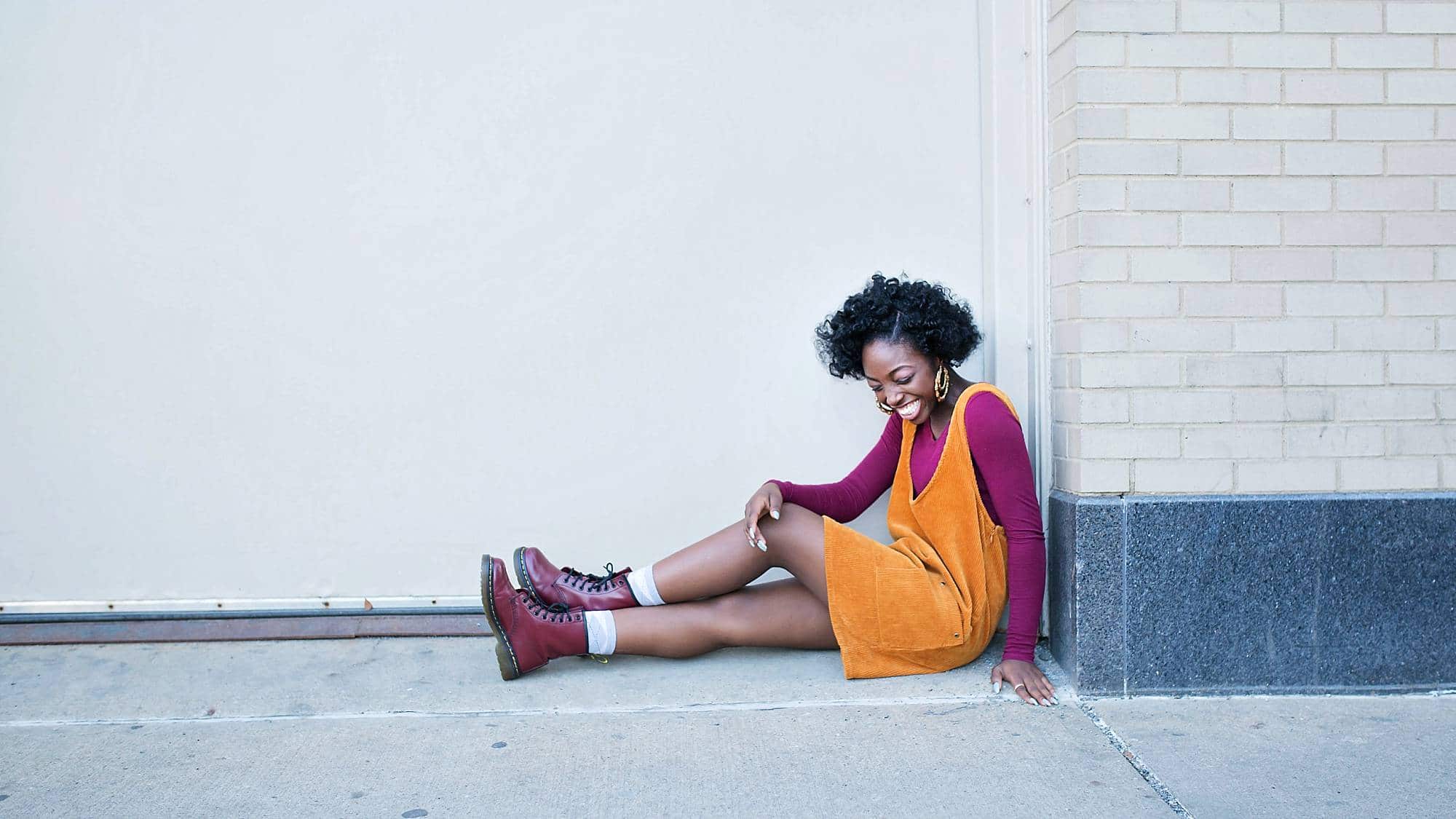 image of senior girl wearing orange jumper, sitting on a city sidewalk, looking down, laughing