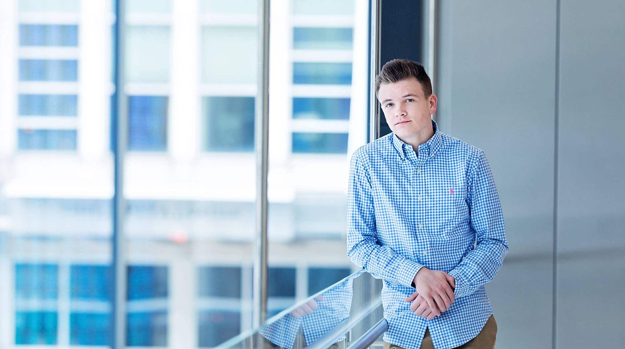 image of a senior guy in light blue button down shirt leaning against a glass railing inside a downtown atrium