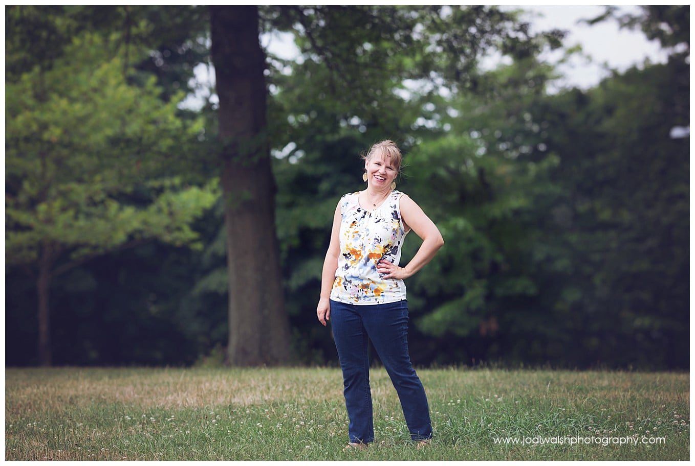 image of a woman in blue jeans and a sleeveless top standing in a grassy field with large trees behind her from a headshot session.