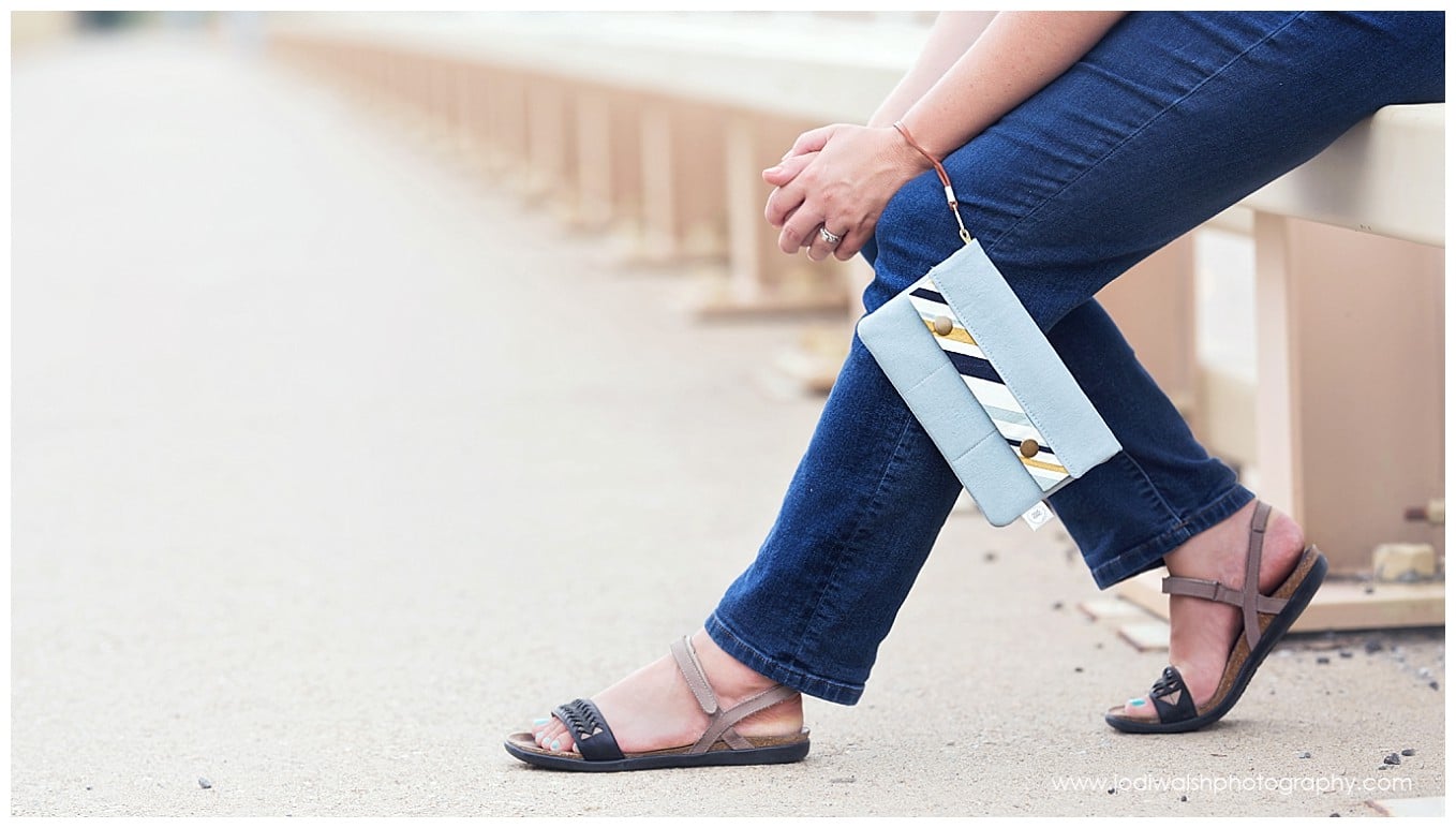 image of legs in blue jeans and sandals. hands are held at the knee and a blue wallet purse hangs from the wrist